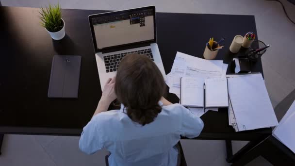 Jung brünette im die büro. Sie sitzt vor dem Laptop-Monitor. Draufsicht. Girle drückt die Tasten der Tastatur. — Stockvideo