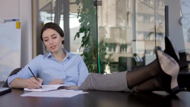 Trabajadora de oficina joven sentada en la mesa. Sus pies sobre la mesa. Ella escribe en el cuaderno. — Vídeos de Stock