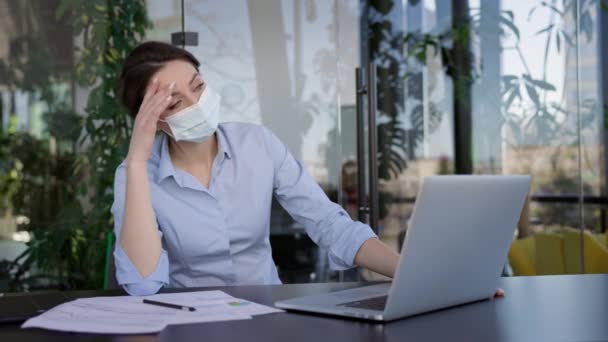 Junges Mädchen im Büro. Sie sitzt mit Maske am Tisch, um sich vor Viren zu schützen. Mädchen arbeiten am Laptop. Sie massiert ihre Tempel. — Stockvideo