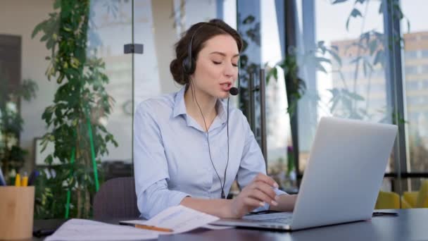 Jovencita en la oficina. Cara de chica joven. Ella está en auriculares y con micrófono. Ella habla y escribe en el teclado del ordenador portátil. — Vídeos de Stock