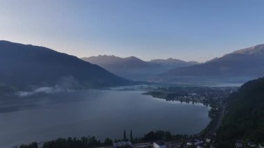 Geniş alp gölü manzarası çevresindeki tepeler ve uzak vadilerle, erken aydınlık yamaçları ve sakin suları boyar.
