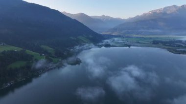Yüzen sis parçalarıyla benekli gölün hava aracı görüntüsü ve ötesindeki berrak vadi, ilk ışık çizgileri kıyı şeridini aydınlatıyor