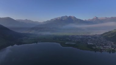 Hava manzarası şafak vakti alp tepeleri, yumuşak mavi ışık, uzak köy kıyı şeridi ve sakin yansıtıcı su ile gölün hatlarını örttü.