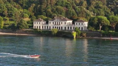 Kristal mavi göl kenarındaki lüks villa, yoğun yeşil ağaçlara yuva yapmış beyaz neoklasik ev, güneş ışığını yansıtan sakin su, kırmızı tekne.