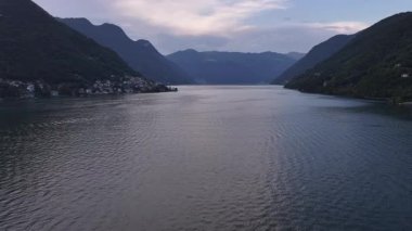 Alacakaranlık gölü sahnesi yumuşak mor gökyüzü ve yumuşak dalgalar, siluetli dağlar durgun suyu çerçeveliyor, sakin akşam havası