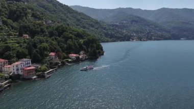 Zümrüt suyuyla göl kenarındaki köye doğru hava yaklaşımı ve yükselen tekneler, dik yeşil yamaçtaki teraslı evler, yaz mevsimi.