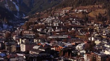 Tepenin yamacında güneşli bir dağ köyü kümelenmiş dağ evleri ve kar yamaçları, dolambaçlı yollar ve kilise kulesi, panoramik insansız hava aracı taraması
