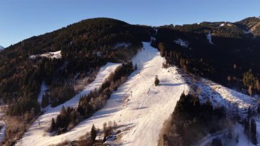 Gün batımında hava karlı alp kayak yamacı, altın ışık, bakımlı koşular boyunca uzun gölgeler bırakıyor, sık ve yeşil ormanlar etrafını sarıyor.