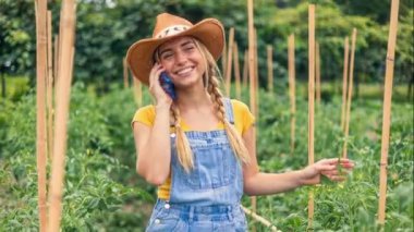 Beyaz kadın çiftçi telefonda domates kazıklarının arasında konuşuyor, hasır şapka ve kot tulumlarla gülümsüyor, ekin planlarını ve pazarı tartışıyor.