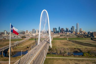 Dallas şehrinin silueti, ikonik Margaret Hunt Hill köprüsü ve Teksas bayrağı dalgalanıyor.