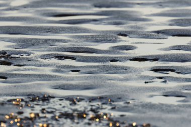 Okyanus sahilindeki güneş ışığının dalgalanmalarını ve yansımalarını gösteren ıslak kum desenleri.