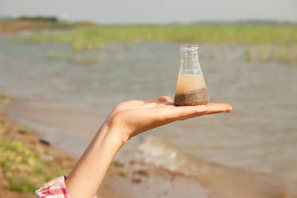 Water Purity test Hand met een chemische kolf of fles met water, meer of rivier op de achtergrond. — Stockfoto