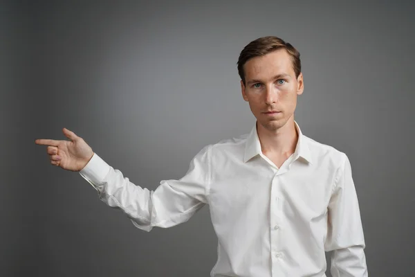 Young man in white shirt is pointing at something, standing on grey background.