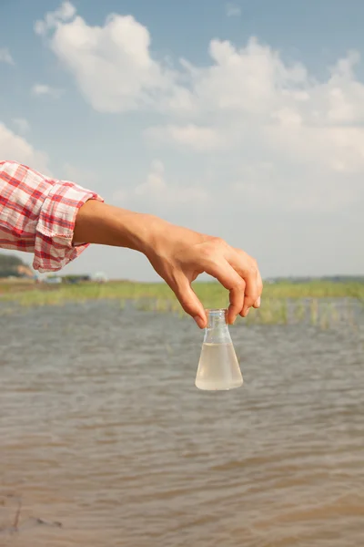 Voda čistoty Test. ruka drží chemická baňka s kapalinou, jezero nebo řeka v pozadí. — Stock fotografie