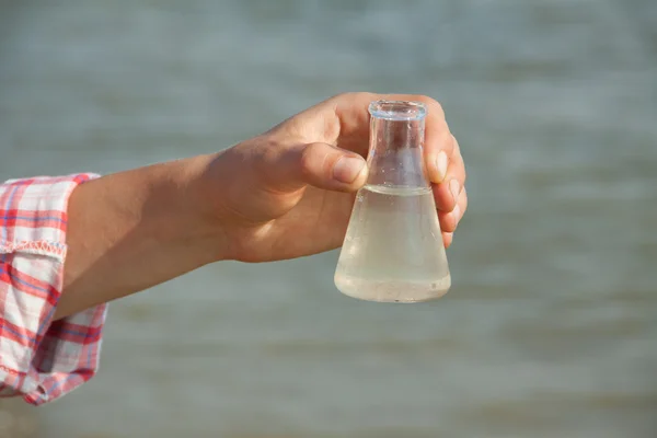 Prueba de pureza del agua. Frasco químico de mano con líquido, lago o río en el fondo . —  Fotos de Stock