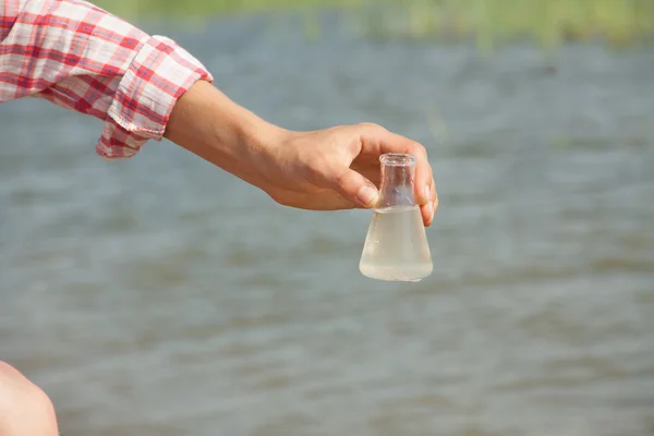 Water Purity test Hand met chemische kolf of fles met vloeistof, meer of rivier op de achtergrond. — Stockfoto