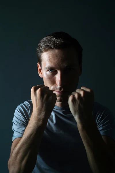 Young man in t-shirt on a dark background holding fists at his face. — Stock Photo, Image