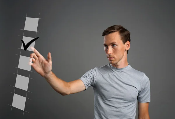 Un joven con camiseta marcando la casilla de la lista. Fondo gris . — Foto de Stock