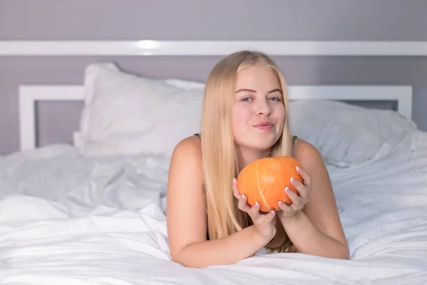 Hermosa rubia feliz con una calabaza en la cama con sábanas blancas. concepto de Halloween — Foto de Stock