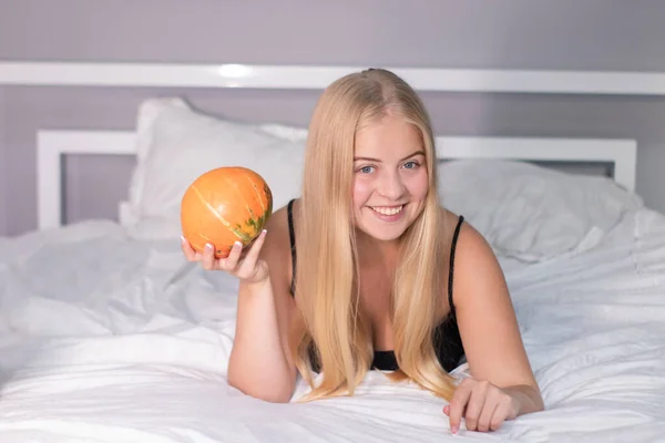 Красивая счастливая блондинка с тыквой на кровати с белыми простынями. Хэллоуин — стоковое фото