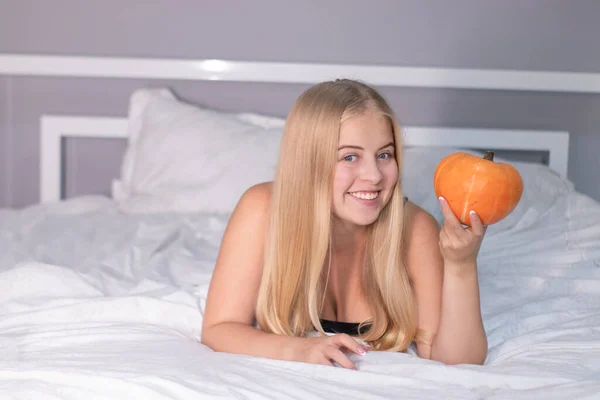 Schöne glückliche Blondine mit einem Kürbis auf dem Bett mit weißen Laken. Halloween-Konzept — Stockfoto