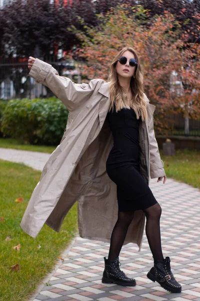 Attractive slim young brunette in fashionable trench, black dress and sunglasses in park. autumn, yellow leaves — Stock Photo, Image