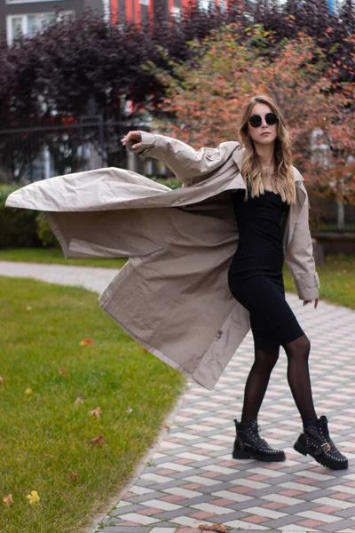 Atraente slim jovem morena na trincheira da moda, vestido preto e óculos de sol no parque. Outono, folhas amarelas — Fotografia de Stock