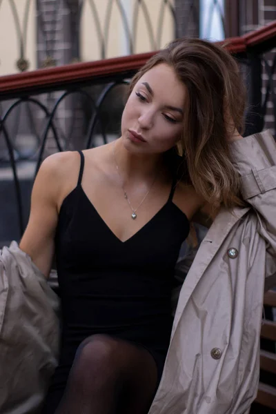 Atractiva morena joven delgada en gabardina de moda y vestido negro sentado en un banco. otoño, hojas amarillas — Foto de Stock