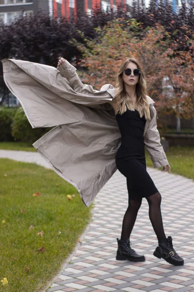 Atraente slim jovem morena na trincheira da moda, vestido preto e óculos de sol no parque. Outono, folhas amarelas — Fotografia de Stock