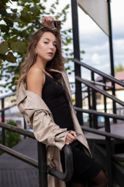 şık trençkotlu ve merdivenlerin yanında yürüyen siyah elbiseli güzel, zayıf, genç bir esmer. Sonbahar, sarı yapraklar.