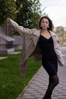 şık trençkotlu ve siyah elbiseli çekici, zayıf, genç bir esmer.