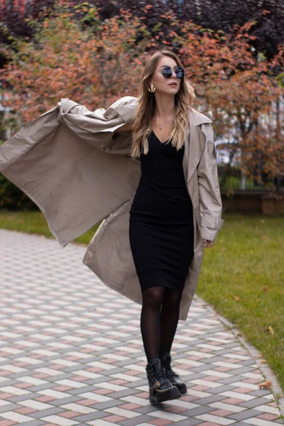 Ελκυστική λεπτή μελαχρινή σε μοντέρνο χαράκωμα, μαύρο φόρεμα και γυαλιά ηλίου στο πάρκο. Φθινόπωρο, κίτρινα φύλλα — Φωτογραφία Αρχείου