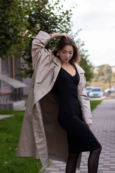 Attractive slim young brunette in fashionable trench coat and black dress — Stock Photo, Image