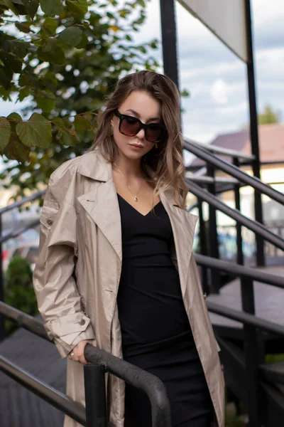 Mooie slanke brunette in modieuze trench jas, zwarte jurk en zonnebril wandelen in de buurt van trappen. herfst gele bladeren — Stockfoto