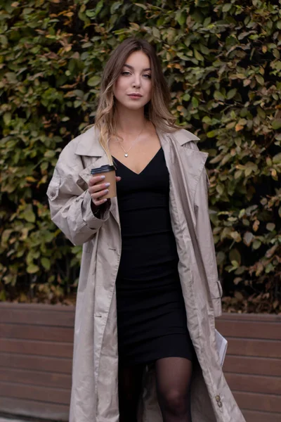 Hermosa delgada joven morena en gabardina de moda y vestido negro con una taza de café de papel — Foto de Stock