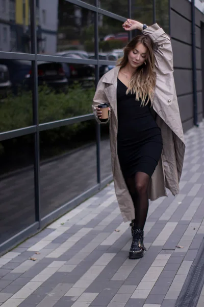Bela jovem morena em casaco de trincheira na moda e vestido preto andando pela rua. Outono, folhas amarelas. — Fotografia de Stock