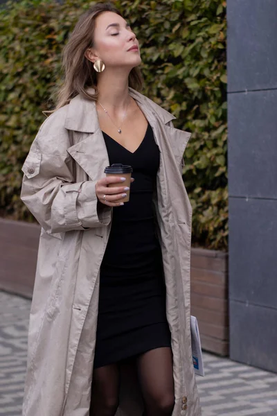 Beautiful young brunette in fashionable trench coat and black dress walking through the street. autumn, yellow leaves. — Stock Photo, Image