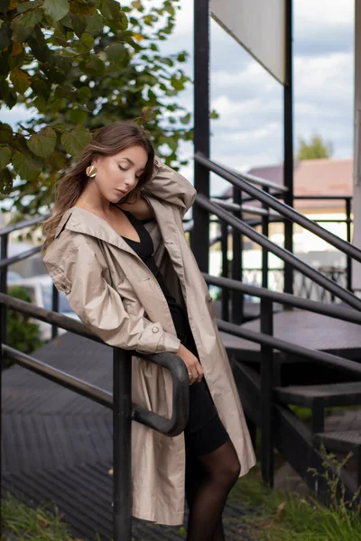 Vacker smal ung brunett i fashionabla trenchcoat och svart klänning promenader nära trappor. höst, gula blad. — Stockfoto