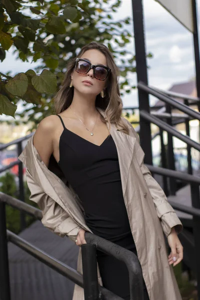 Mooie slanke brunette in modieuze trench jas, zwarte jurk en zonnebril wandelen in de buurt van trappen. herfst gele bladeren — Stockfoto