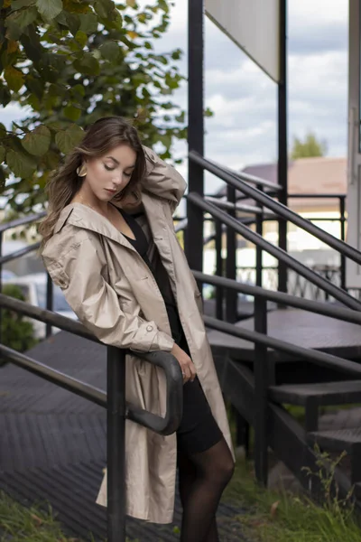 Belle jeune brune mince en trench-coat à la mode et robe noire marchant près des escaliers. automne, feuilles jaunes. — Photo