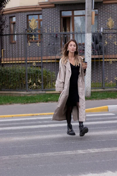 Hermosa delgada joven morena en gabardina de moda y vestido negro cruzan un cruce peatonal — Foto de Stock