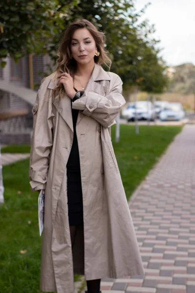 Attractive slim young brunette in fashionable trench coat and black dress with a newspaper — Stock Photo, Image