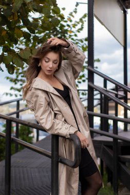 şık trençkotlu ve merdivenlerin yanında yürüyen siyah elbiseli güzel, zayıf, genç bir esmer. Sonbahar, sarı yapraklar.