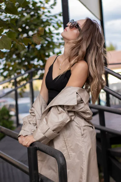 Mooie slanke brunette in modieuze trench jas, zwarte jurk en zonnebril wandelen in de buurt van trappen. herfst gele bladeren — Stockfoto