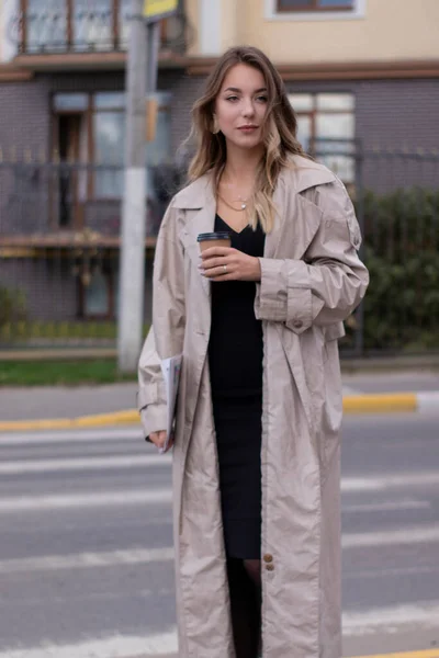 Beautiful slim young brunette in fashionable trench coat and black dress cross a pedestrian crossing — Stock Photo, Image