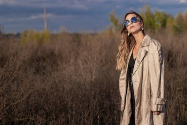 Modaya uygun trençkotlu esmer, siyah elbiseli ve güneş gözlüklü. Sonbahar, sarı yapraklar, mavi gökyüzü