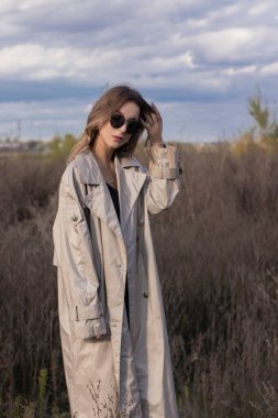 Modaya uygun trençkotlu esmer, siyah elbiseli ve güneş gözlüklü. Sonbahar, sarı yapraklar, mavi gökyüzü