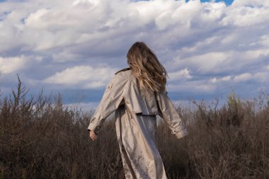 Modaya uygun trençkotlu esmer, siyah elbiseli ve güneş gözlüklü. Sonbahar, sarı yapraklar, mavi gökyüzü