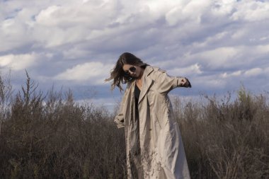 Modaya uygun trençkotlu esmer, siyah elbiseli ve güneş gözlüklü. Sonbahar, sarı yapraklar, mavi gökyüzü