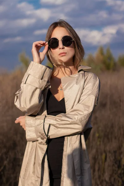 Brunette in modieuze trench jas, zwarte jurk en zonnebril in het veld. herfst, gele bladeren, blauwe lucht — Stockfoto